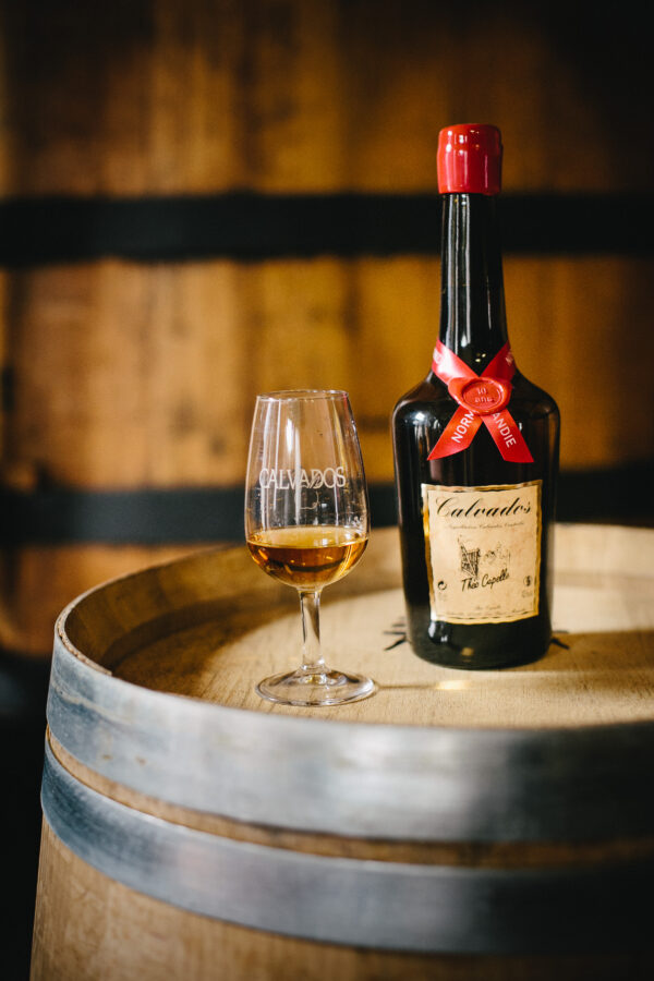 Bouteille de Calvados et verre sur un tonneau en bois, présentant un produit de la Cidrerie-Distillerie Théo Capelle.