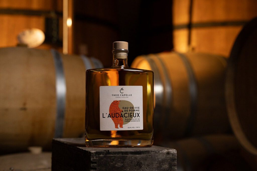 Bouteille d'eau-de-vie de pomme "L'Audacieux" de la Cidrerie-Distillerie Théo Capelle, posée sur un cube près des fûts en bois.