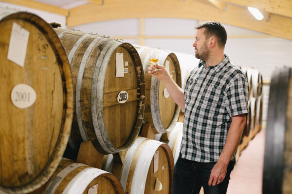 Homme observant un verre de Calvados AOC 5 ans dans une cidrerie, entouré de fûts en bois.
