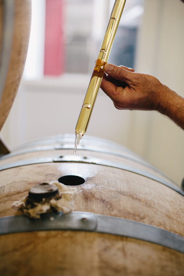 Main tenant un échantillon de liquide doré au-dessus d'un tonneau en bois, symbolisant le processus de production de Calvados.