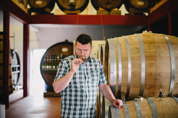 Homme en chemise à carreaux dégustant un Calvados dans une distillerie, entouré de fûts en bois.
