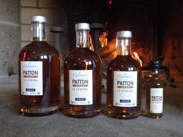 Bouteilles de Calvados "Patton" en différentes tailles, devant une cheminée, mettant en valeur le savoir-faire de la Cidrerie-Distillerie Théo Capelle.