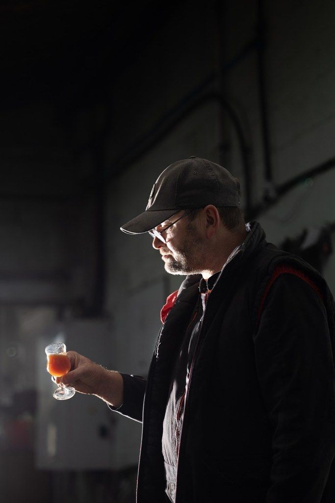 Un artisan de la cidrerie Théo Capelle observe un verre de cidre dans un atelier, lumière tamisée.