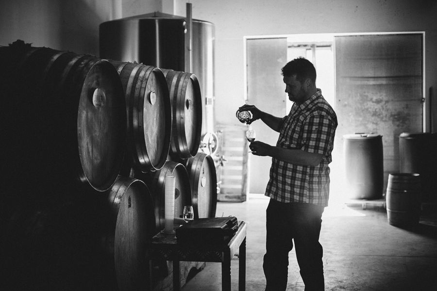 Homme en train de verser une boisson dans un verre, entouré de fûts dans une distillerie.