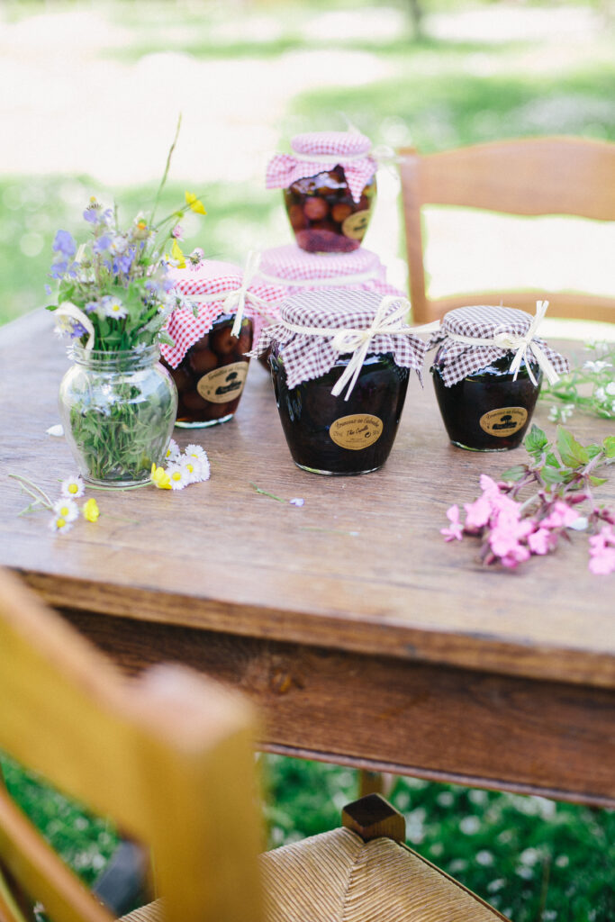 Bocaux de griottes au Calvados, décorés de tissu à carreaux, sur une table en bois avec des fleurs sauvages.