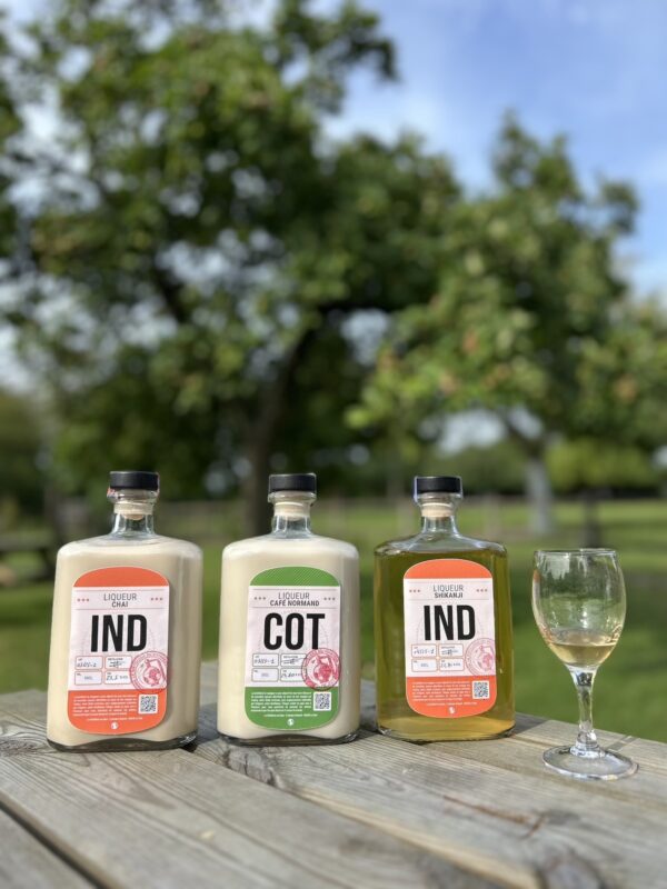 Trois bouteilles de liqueurs artisanales de la Cidrerie-Distillerie Théo Capelle, avec un verre et un décor verdoyant en arrière-plan.