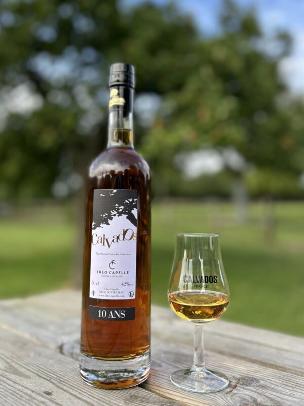 Bouteille de Calvados AOC 10 ans et verre, produit par la cidrerie Théo Capelle, en arrière-plan d'un paysage normand.