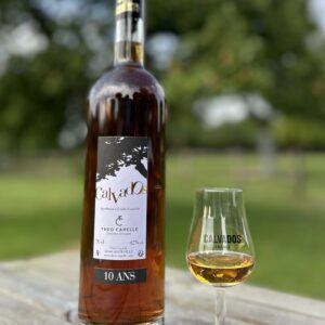 Bouteille de Calvados AOC 10 ans et verre, présentés sur une table en bois, en arrière-plan flou de vergers.