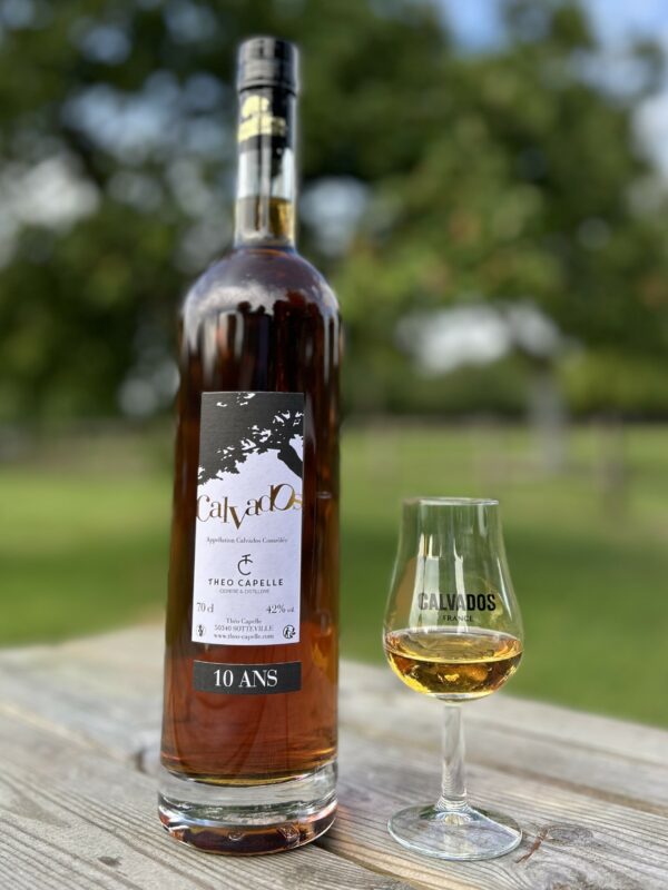 Bouteille de Calvados AOC 10 ans et verre, présentés sur une table en bois, en arrière-plan flou de vergers.