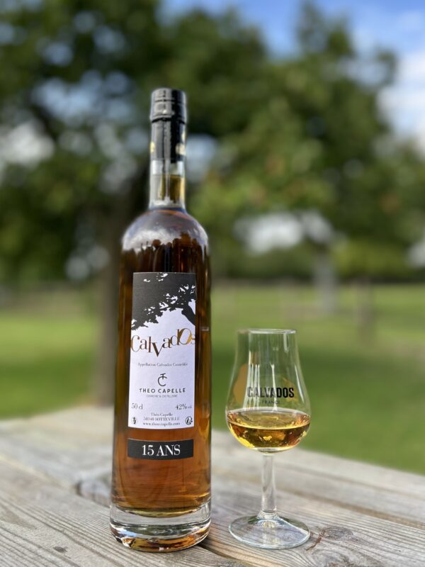 Bouteille de Calvados AOC 15 ans de 50 cl et verre, en arrière-plan, verger normand.