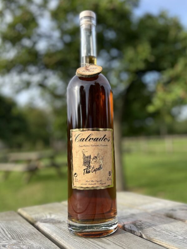 Bouteille de Calvados de la Cidrerie-Distillerie Théo Capelle, sur une table en bois, avec un fond d'arbres.