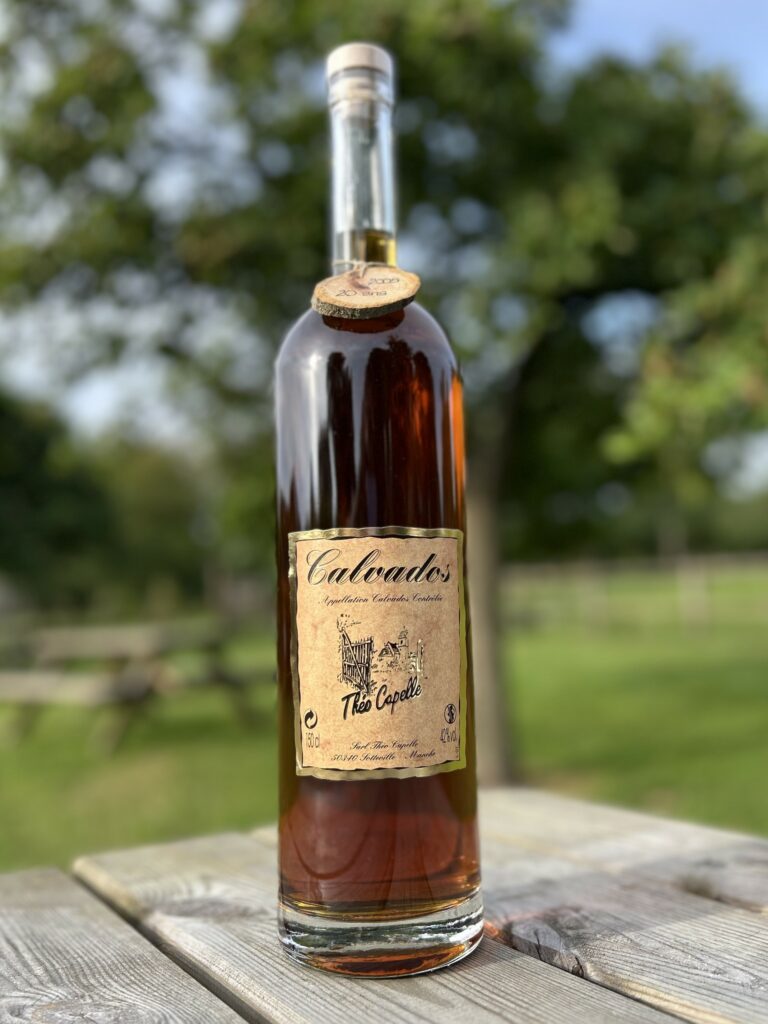 Bouteille de Calvados de la Cidrerie-Distillerie Théo Capelle, sur une table en bois, avec un fond d'arbres.