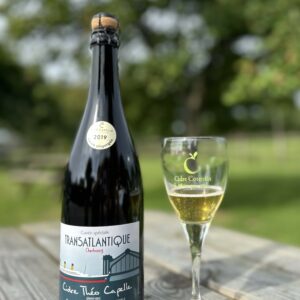 Bouteille de cidre demi-sec Transatlantique de la cidrerie Théo Capelle, accompagnée d'un verre sur une table en bois.