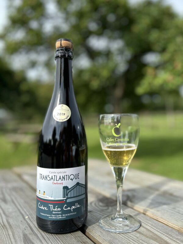 Bouteille de cidre demi-sec Transatlantique de la cidrerie Théo Capelle, accompagnée d'un verre sur une table en bois.