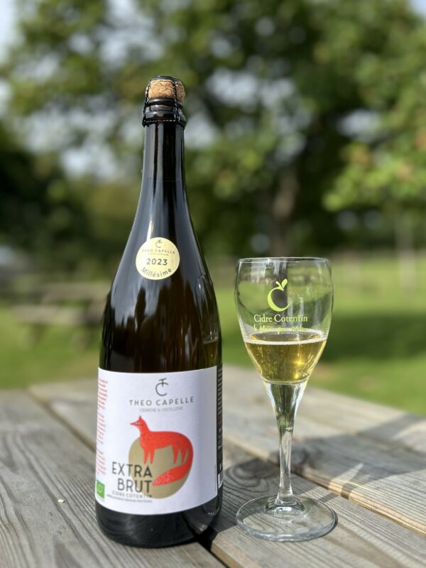 Bouteille de cidre AOP Cotentin Extra Brut 2023 avec un verre, sur une table en bois dans un cadre naturel.