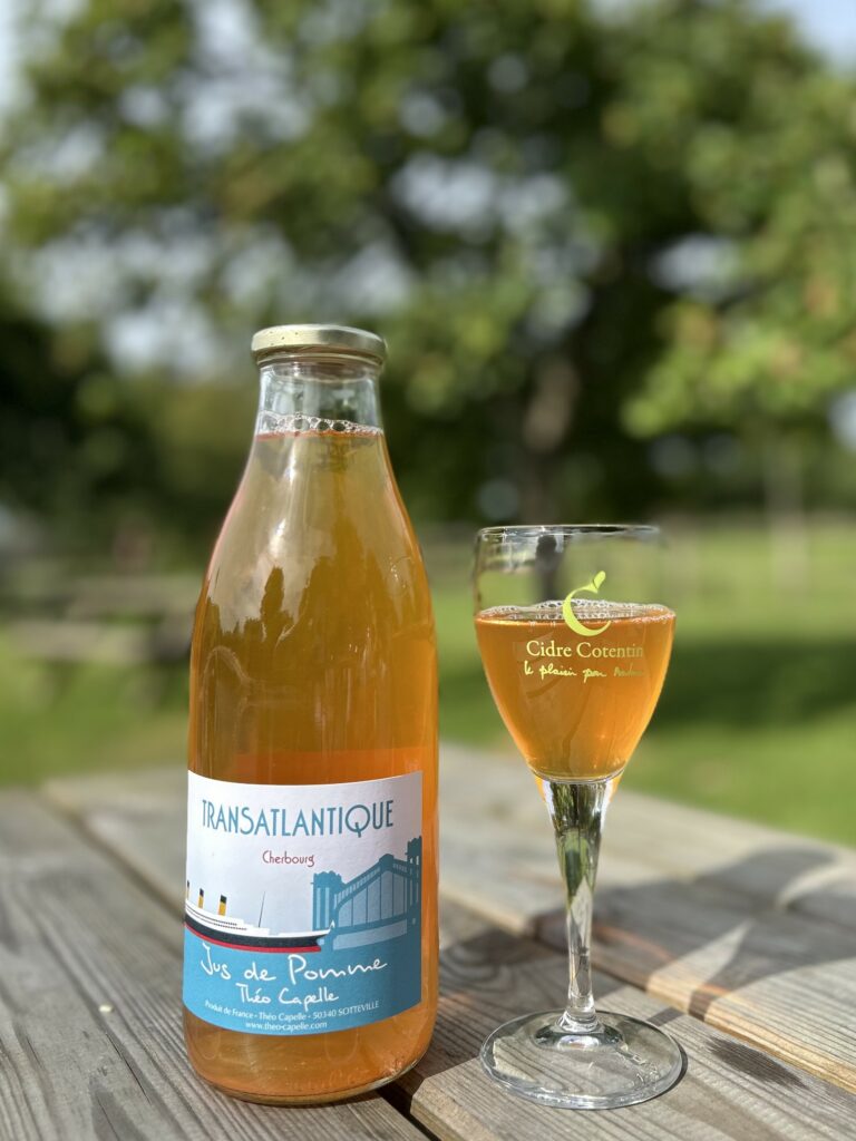 Bouteille de jus de pomme acidulé Théo Capelle, servie dans un verre sur une table en bois, en arrière-plan un décor verdoyant.