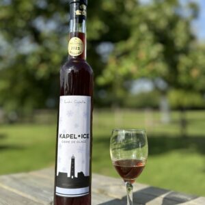 Bouteille de Kapel Ice, cidre de glace, avec un verre sur une table en bois, en arrière-plan un paysage verdoyant.
