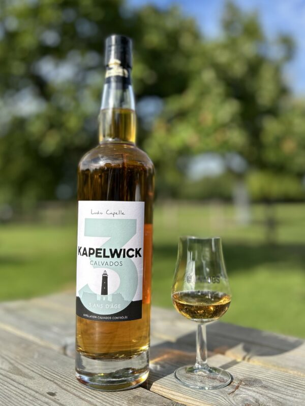 Bouteille de Calvados KAPELWICK 3 ans d'âge, accompagnée d'un verre, sur une table en bois en extérieur.