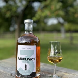 Bouteille de Calvados 3 ans d’âge Kapelwick et un verre, posée sur une table en bois, avec un paysage verdoyant en arrière-plan.