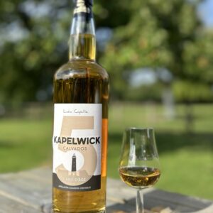 Bouteille de Calvados AOC 5 ans d'âge, accompagnée d'un verre, sur une table en extérieur, avec un paysage verdoyant en arrière-plan.