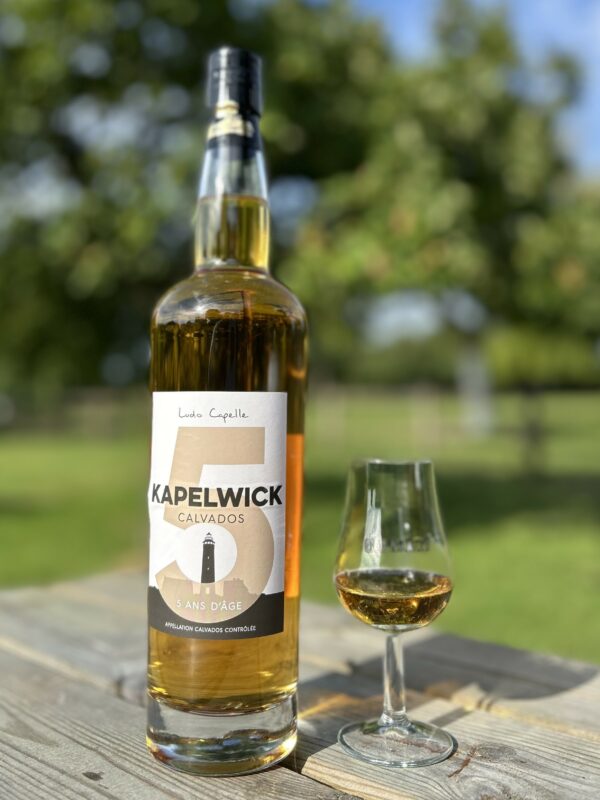 Bouteille de Calvados AOC 5 ans d'âge, accompagnée d'un verre, sur une table en extérieur, avec un paysage verdoyant en arrière-plan.