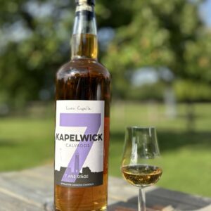 Bouteille de Calvados KAPELWICK 7 ans d'âge avec un verre, sur une table en bois, en arrière-plan de vergers.