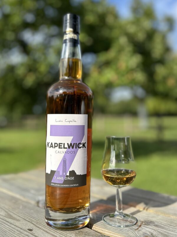 Bouteille de Calvados KAPELWICK 7 ans d'âge avec un verre, sur une table en bois, en arrière-plan de vergers.