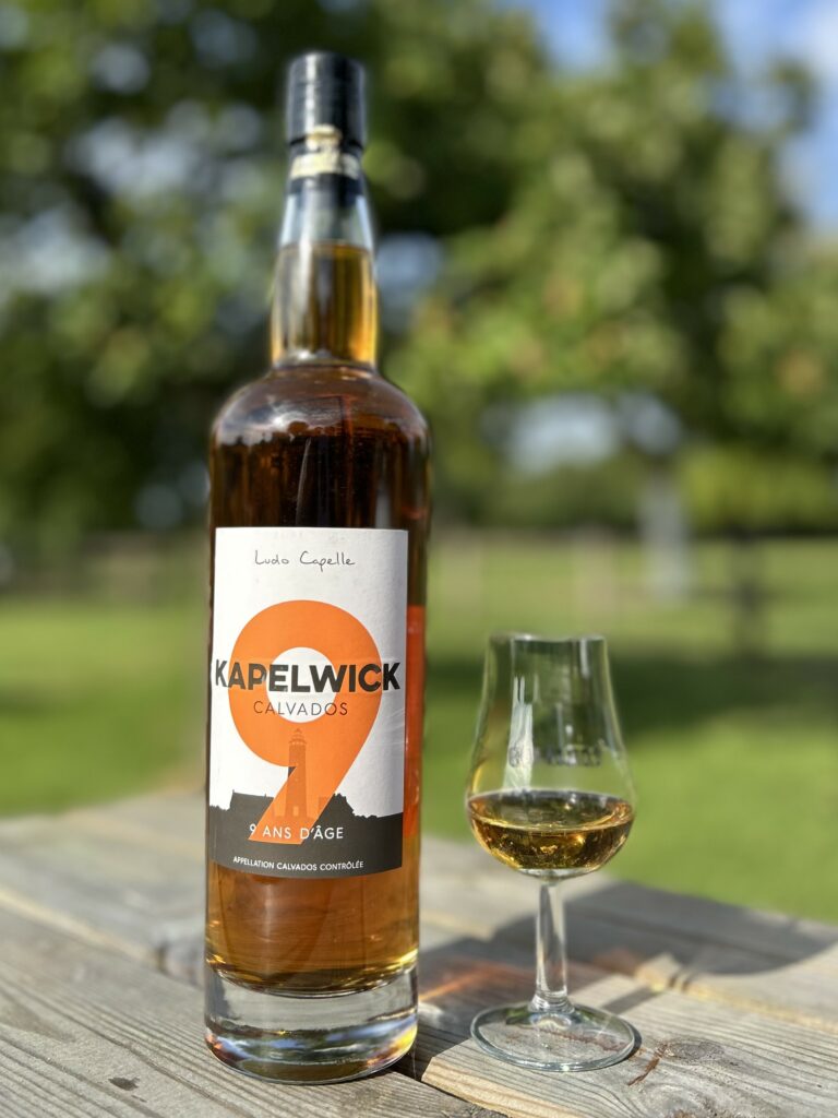 Bouteille de Calvados KAPELWICK 9 ans et un verre sur une table en bois, entourée de verdure.
