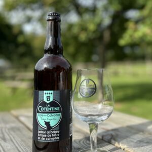 Bouteille de bière "La Cotentine" 75cl et un verre, sur une table en bois, entourés de verdure.