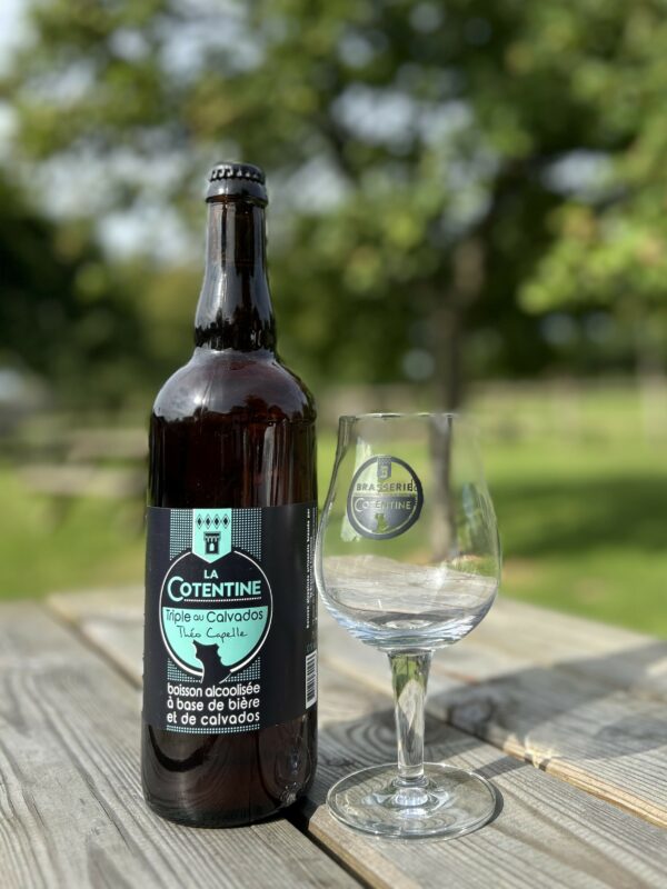 Bouteille de bière "La Cotentine" 75cl et un verre, sur une table en bois, entourés de verdure.