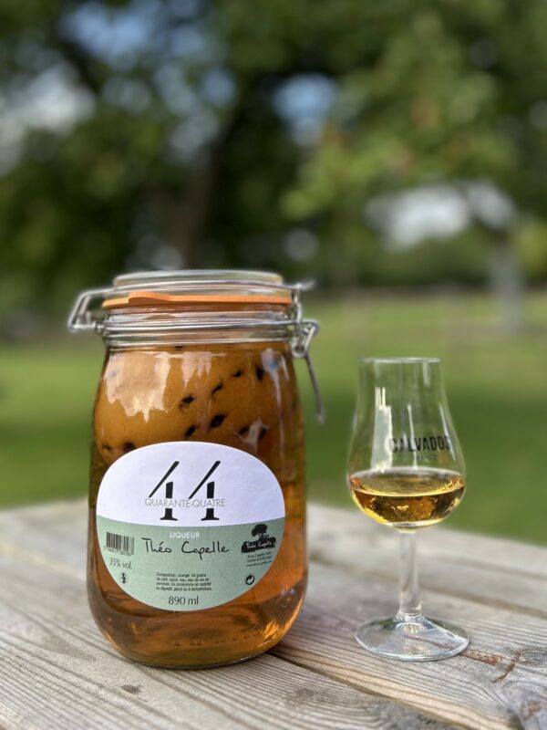 Bocal de liqueur 44 Quarante-Quatre de Théo Capelle avec un verre de Calvados sur une table en bois, entouré de verdure.
