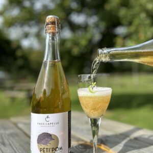 Bouteille de jus de pomme pétillant biologique PÉT'POM de la Cidrerie Théo Capelle, versée dans un verre, en pleine nature.