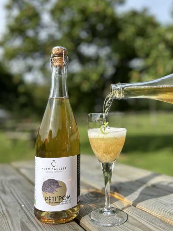Bouteille de jus de pomme pétillant biologique PÉT'POM de la Cidrerie Théo Capelle, versée dans un verre, en pleine nature.