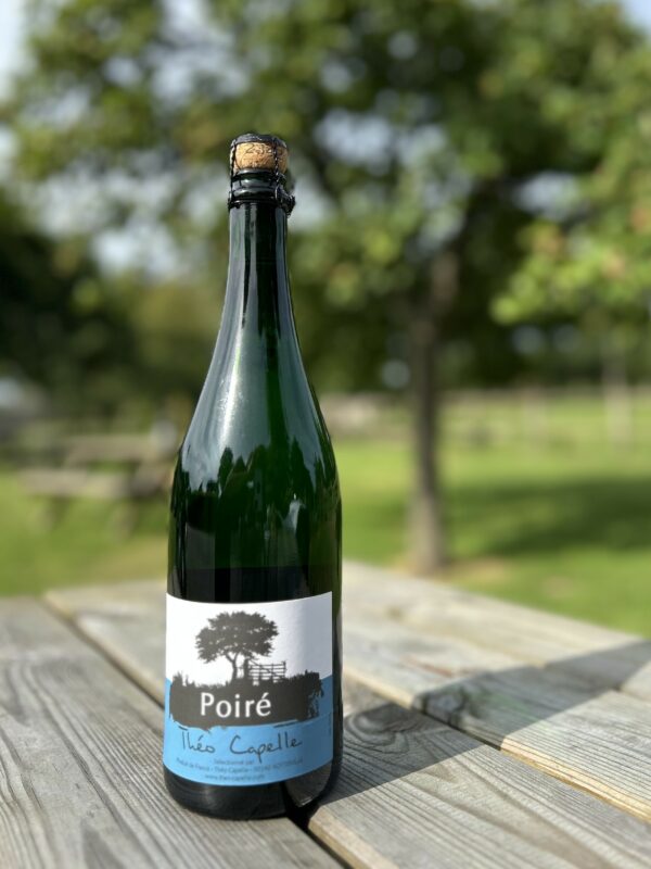 Bouteille de Poiré de la Cidrerie-Distillerie Théo Capelle sur une table en bois, en arrière-plan flou de verdure.